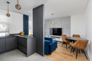 Na zdjęciu widać kolejny przykład realizacji firmy Pixel Kuchnie, która specjalizuje się w projektowaniu mebli kuchennych na indywidualne zamówienie. Centralnym punktem jest nowoczesna kuchnia z eleganckimi szaro-białymi frontami oraz czarnym blatem, która została wykonana na wymiar, zapewniając maksymalne wykorzystanie dostępnej przestrzeni. Wysoka jakość wykonania mebli oraz zastosowanie trwałych materiałów podkreślają luksusowy charakter wnętrza. W tle widoczny jest salon z komfortowym niebieskim meblem wypoczynkowym, który świetnie komponuje się z ciepłym odcieniem drewnianego parkietu. Nad stołem jadalnym wiszą designerskie, brązowe krzesła, a nad nimi zawieszono nowoczesną lampę w złotym kolorze, co razem tworzy spójną i elegancką całość. Przestronność i otwarty układ kuchni z salonem są przykładem na to, jak firma Pixel Kuchnie potrafi dostosować swoje projekty do współczesnych trendów w aranżacji wnętrz, jednocześnie zachowując unikalny i personalizowany charakter każdego zamówienia.