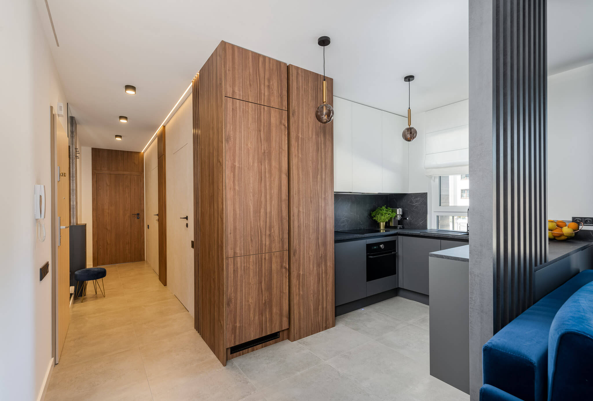 Zdjęcie przedstawia nowoczesną kuchnię zaprojektowaną na wymiar przez firmę Pixel Kuchnie. Widoczna jest elegancka zabudowa kuchenna z frontami w odcieniach szarości i bieli, która wydaje się idealnie dopasowana do przestrzeni, oferując zarówno styl, jak i funkcjonalność. Meble kuchenne z ciemnego drewna, które harmonizują z minimalistycznym designem, wskazują na możliwość personalizacji według potrzeb klienta. Całość jest dobrze oświetlona dzięki zastosowaniu dyskretnego oświetlenia LED, które podkreśla nowoczesny charakter kuchni. Dodatki takie jak elegancka, ciemna blacha marmurowa oraz wiszące, szklane lampy dopełniają wyrafinowany wygląd. Kuchnia wyposażona jest w nowoczesne urządzenia AGD, co jest dowodem na to, że Pixel Kuchnie dba o to, aby ich kuchnie były nie tylko piękne, ale i praktyczne.